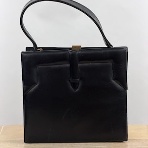 May include: A structured black handbag with a single top handle and a rectangular silhouette. The front features a central vertical strap detail and a metallic gold-toned clasp closure at the top center. The surface has a smooth, matte finish.
