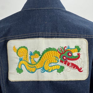May include: A dark blue denim jacket with a white patch on the back featuring an embroidered yellow dragon with green and red accents.