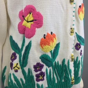 May include: White knitted cardigan with embroidered flowers and bees. The flowers are pink, yellow, orange, and purple. The bees are yellow and black.