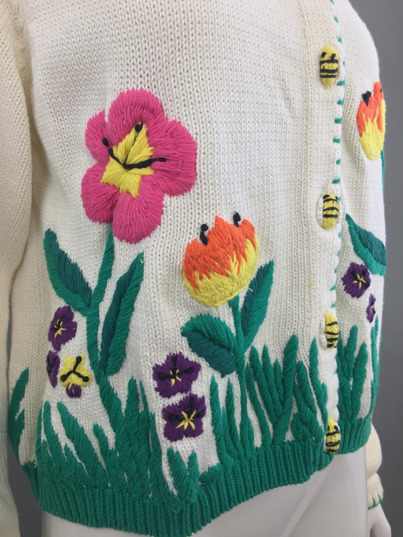 May include: White knitted cardigan with embroidered flowers and bees. The flowers are pink, yellow, orange, and purple. The bees are yellow and black.