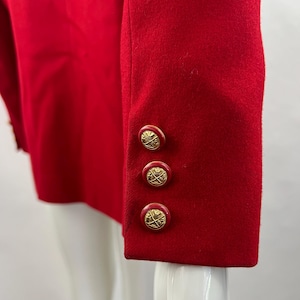 May include: Close-up of a red blazer sleeve with three gold-colored buttons. The buttons have a decorative design. The blazer is made of a smooth fabric and is displayed on a mannequin.