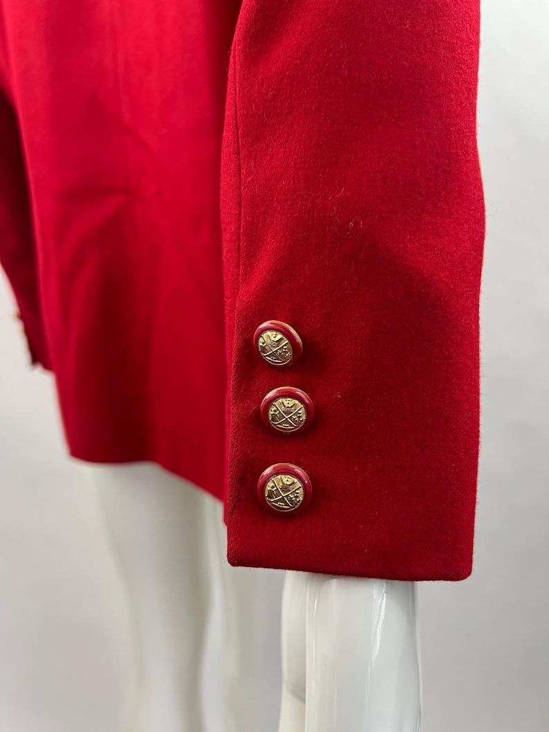 May include: Close-up of a red blazer sleeve with three gold-colored buttons. The buttons have a decorative design. The blazer is made of a smooth fabric and is displayed on a mannequin.