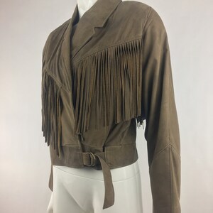 May include: A brown suede jacket with fringe detailing and a belt. The jacket has a long sleeve and a cropped length.
