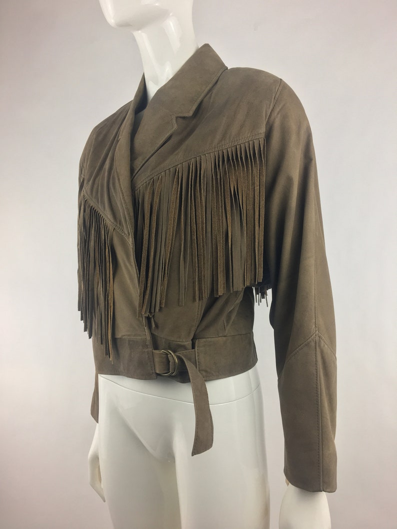 May include: A brown suede jacket with fringe detailing and a belt. The jacket has a long sleeve and a cropped length.