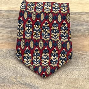 May include: A necktie featuring a deep red background with a repeating pattern of blue and yellow floral motifs arranged in vertical columns, interspersed with small beige and dark blue diamond-shaped geometric accents throughout the fabric.