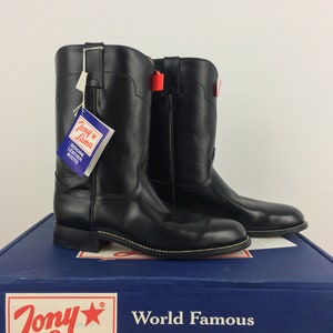 May include: A pair of black leather cowboy boots with a pointed toe and a low heel. The boots are sitting on a blue box with the words "Tony Lama World Famous Handcrafted Boots" printed on it.