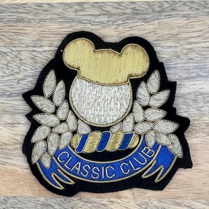 May include: A black felt patch with a gold and silver embroidered design. The design features a bear head with a gold hat, surrounded by silver leaves and a blue ribbon with the text "CLASSIC CLUB".