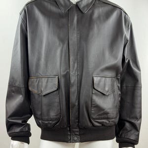 Può includere: Una giacca in pelle marrone scuro con colletto classico, maniche lunghe con polsini a coste e orlo a coste. La parte anteriore presenta una chiusura centrale con zip coperta da una patta e due ampie tasche applicate con patte a punta.