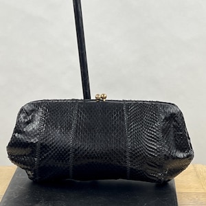 May include: A black clutch bag with a textured snakeskin pattern and a gold-toned metal kiss-lock clasp closure. The rectangular bag features a structured frame and a glossy finish, showcasing a detailed scale texture across the entire exterior surface.