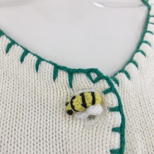 May include: White knitted cardigan with green trim and a bee-shaped button. The cardigan has a V-neck and is made of a soft, lightweight fabric.