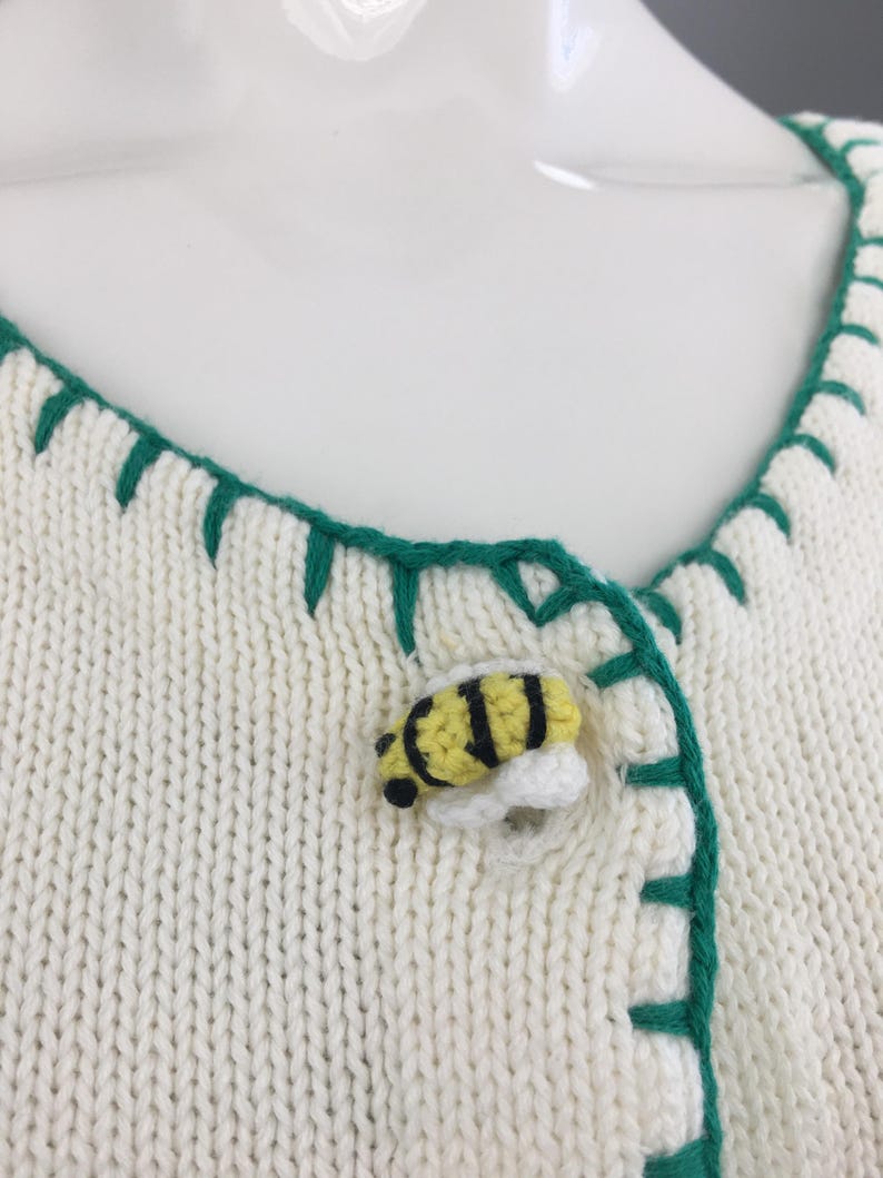 May include: White knitted cardigan with green trim and a bee-shaped button. The cardigan has a V-neck and is made of a soft, lightweight fabric.