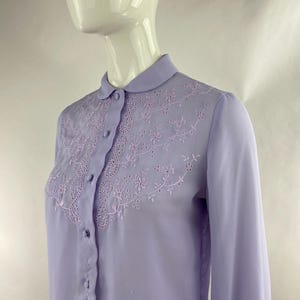 May include: A pale lilac blouse with a rounded collar and long sleeves. The blouse has floral embroidery across the chest and button fastenings down the front. The fabric is lightweight and semi-transparent. The blouse is displayed on a mannequin.