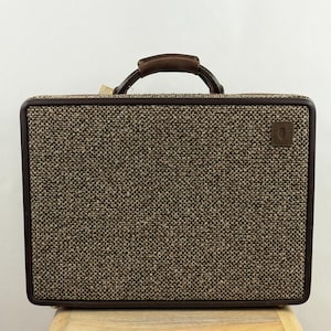 May include: A rectangular briefcase featuring a textured brown and beige tweed fabric exterior. The case is framed by a dark brown border and includes a matching dark brown top handle. A small, square brown leather patch is located on the upper right.