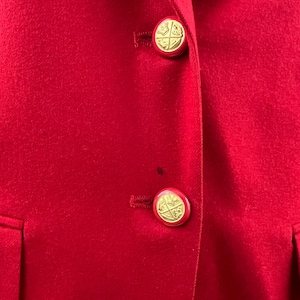 May include: Close-up of a red blazer with two gold-colored buttons. The buttons have a circular design with a gold emblem. The blazer has a classic design with a notched lapel and two front pockets.