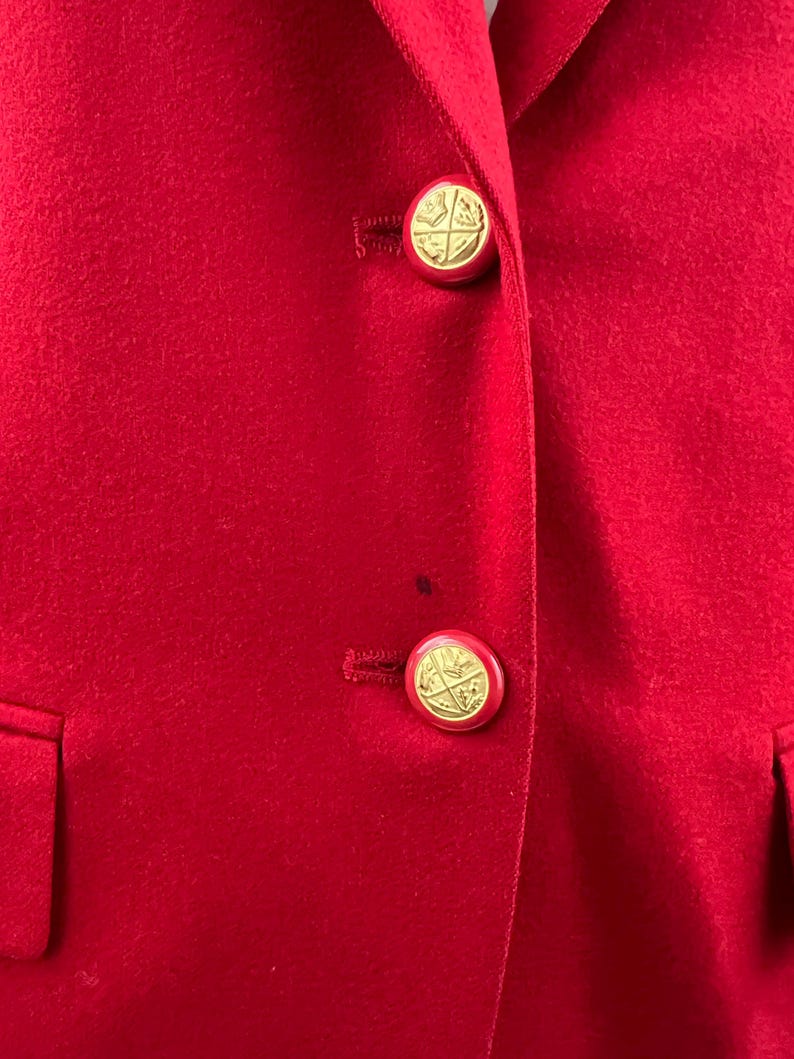 May include: Close-up of a red blazer with two gold-colored buttons. The buttons have a circular design with a gold emblem. The blazer has a classic design with a notched lapel and two front pockets.