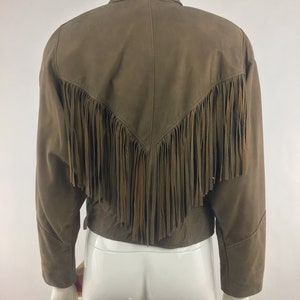 May include: A brown suede jacket with fringe detailing on the back. The jacket has a collared neckline and long sleeves.