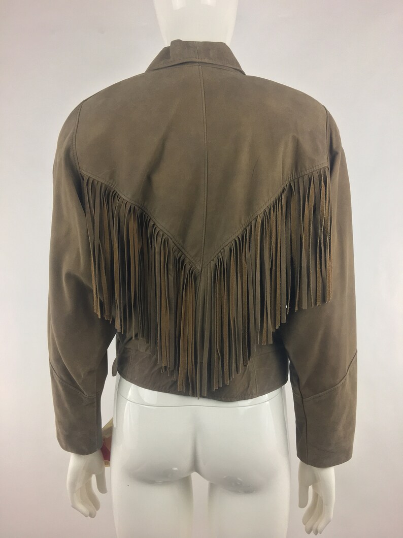 May include: A brown suede jacket with fringe detailing on the back. The jacket has a collared neckline and long sleeves.