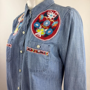 May include: A light blue button-down shirt featuring a large circular red patch on the chest with colorful floral embroidery. The shirt includes two chest pockets, each accented with a horizontal band of intricate, multi-colored geometric embroidery.
