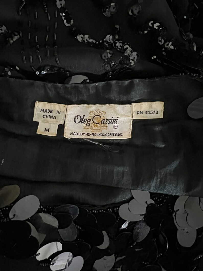 Op de afbeelding: Zwart top met pailletten met een label met de tekst "Oleg Cassini" en "Made in China" in maat "M" en RN 62313.