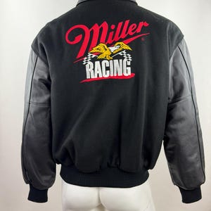 Może przedstawiać: Czarna kurtka typu varsity z długimi czarnymi rękawami i czarnym kołnierzem. Z tyłu znajduje się wyhaftowane logo z czerwonym napisem Miller, grafiką żółtego orła oraz białym napisem RACING nad dwiema szachownicami na czarnym tle.