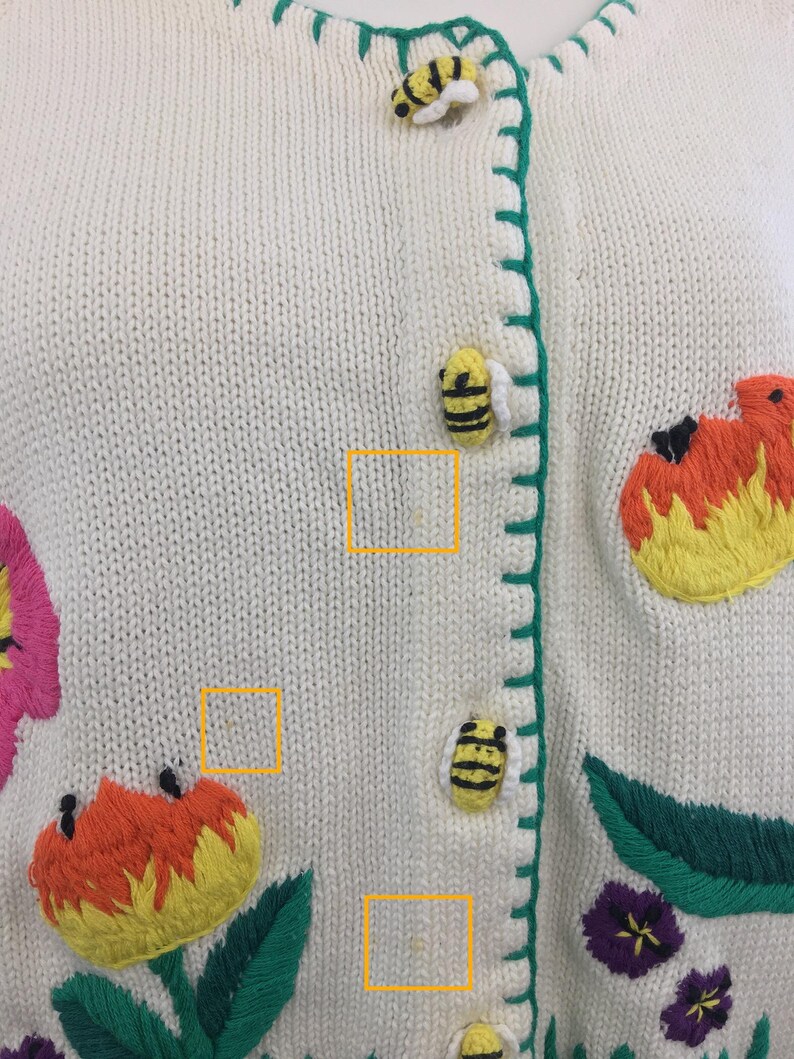 May include: A white knitted cardigan with green trim and embroidered flowers and bees. The cardigan has a button closure and is made of a soft, comfortable material.