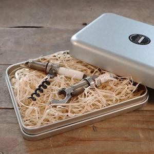 Può includere: Una scatola di metallo argentata con un logo nero sul coperchio. La scatola è aperta e contiene due candele, un cavatappi e trucioli di legno.