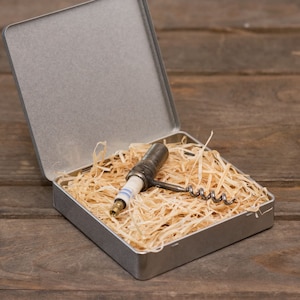 Può includere: Una scatola di metallo argentata con il coperchio aperto, che rivela un cavatappi vintage con manico in legno, annidato in carta triturata.