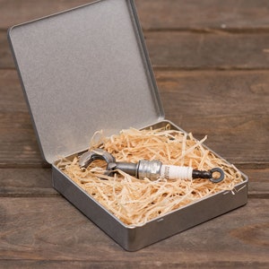 Può includere: Una scatola di metallo argentata con una candela di accensione all'interno. La scatola è piena di carta triturata e la candela di accensione è appoggiata sulla carta.