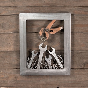 May include: Metal artwork featuring a rectangular frame with a collection of tools. Inside the frame, there are several wrenches and nails, with a pair of orange pliers holding a nut above the wrenches. The background is a wooden surface.