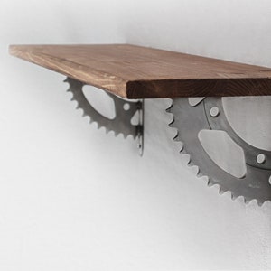 Könnte beinhalten: Ein Holzregal mit Metallkonsolen, die mit Fahrradkettenblättern verziert sind. Das Regal ist aus Naturholz gefertigt und hat einen rustikalen Look.