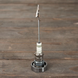May include: A silver metal photo clip holder made from a spark plug, a ball bearing, and a wire. The clip is attached to the wire.