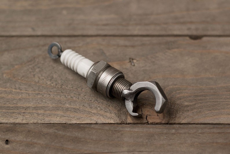 May include: A silver bottle opener made from a repurposed spark plug. The opener has a ring on the top and a C-shaped opening for opening bottles.