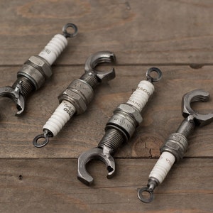 May include: Four silver bottle openers made from repurposed spark plugs. Each opener has a unique design and features a white ceramic insulator with text.