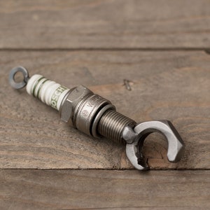 May include: A silver spark plug keychain with a wrench-shaped bottle opener attached. The spark plug is labeled "BOSCH" and has a white ceramic insulator with green markings.