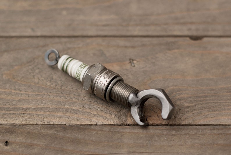 May include: A silver spark plug keychain with a wrench-shaped bottle opener attached. The spark plug is labeled "BOSCH" and has a white ceramic insulator with green markings.