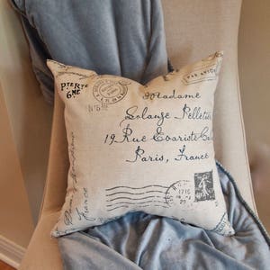 May include: A white pillow with black text that reads "Madame Solange Pelletier 19 Rue Coariste b Paris, France" and a vintage postage stamp design. The pillow is on a light beige chair with a light blue throw blanket.