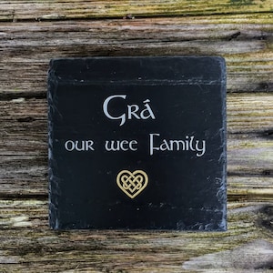 Puede incluir: Un letrero de pizarra negra con las palabras "Grá our wee Family" en letras blancas y un diseño de corazón de nudo celta dorado.