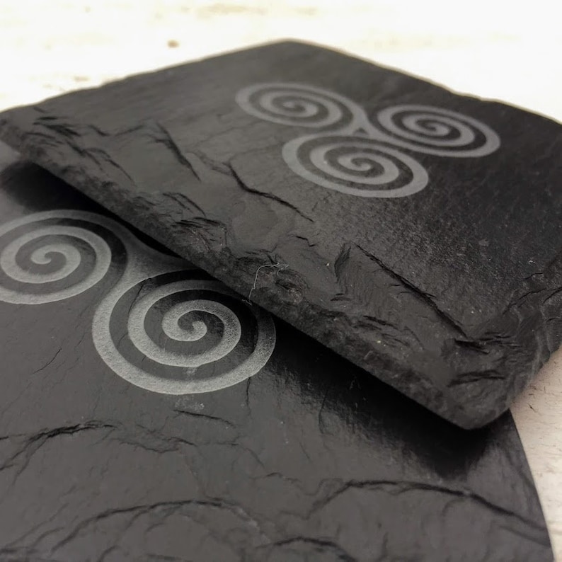 May include: Two black slate coasters with a Celtic knot design. The coasters are made of natural slate and have a rustic look.