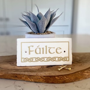 May include: White rectangular sign with the word "Fáilte" in gold lettering. The sign has a Celtic knot design at the bottom and is sitting on a wooden surface. A small succulent plant is in the background.