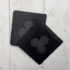 May include: Two black slate coasters with a white Celtic knot design. The coasters are square shaped and have a rough, textured surface.
