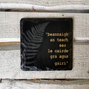 May include: Black slate coaster with a fern design and a gold inscription in Gaelic that reads "beannaigh an teach seo le cairde grá agus gáirí".