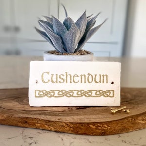 May include: White rectangular sign with the name "Cushendoun" written in gold lettering. The sign has a gold Celtic knot design at the bottom. The sign is sitting on a wooden surface with two gold screws.
