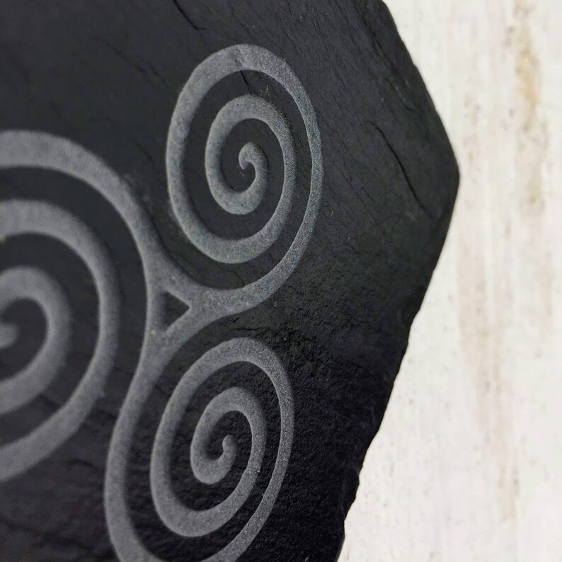 May include: A black slate coaster with a triple spiral design etched into the surface.