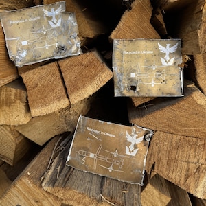 Puede incluir: Tres placas metálicas rectangulares con el texto "Recycled in Ukraine" y un gráfico blanco de un pájaro y un avión se muestran sobre una pila de madera. Las placas tienen un aspecto envejecido y desgastado.
