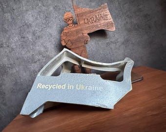 May include: A gray metal object with the words "Recycled in Ukraine" printed on it. A wooden cutout of a person holding a flag with the word "UKRAINE" printed on it is behind the metal object.