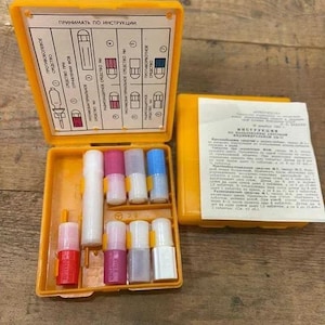 Pode incluir: Um kit de primeiros socorros de plástico amarelo com uma variedade de pequenos frascos e ampolas. O kit tem um rótulo que diz "ПРИНИМАТЬ ПО ИНСТРУКЦИИ", que se traduz como "Tomar de acordo com as instruções".