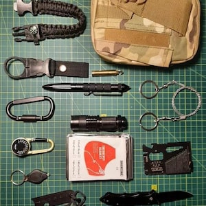Puede incluir: Un conjunto de artículos de equipo de supervivencia, que incluye una bolsa de camuflaje, una linterna, una brújula, un encendedor, una multiherramienta, un brazalete de paracord, un silbato, un cuchillo pequeño y un botiquín de primeros auxilios. El kit está etiquetado como "Emergency Blanket".