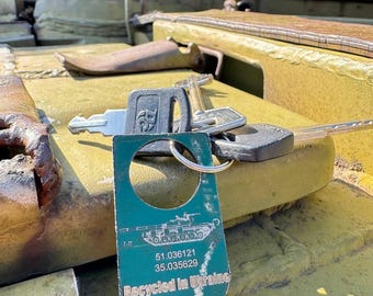 May include: A green tag with a tank silhouette, the text "Recycled in Ukraine", and the coordinates 51.036121 35.035629. The tag is attached to a set of keys on a rusty metal surface.