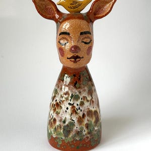 May include: A ceramic figurine featuring a stylised head with deer-like ears and a small yellow bird perched on top. The face has closed eyes, pink cheeks, and a dark mouth. The base has a mottled glaze of brown, white, and green speckles.