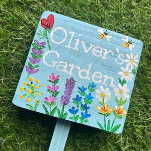 May include: A light blue wooden garden sign with the words "Oliver's Garden" in white. The sign is decorated with painted flowers, bees, and a red poppy. The sign is on a wooden stake.